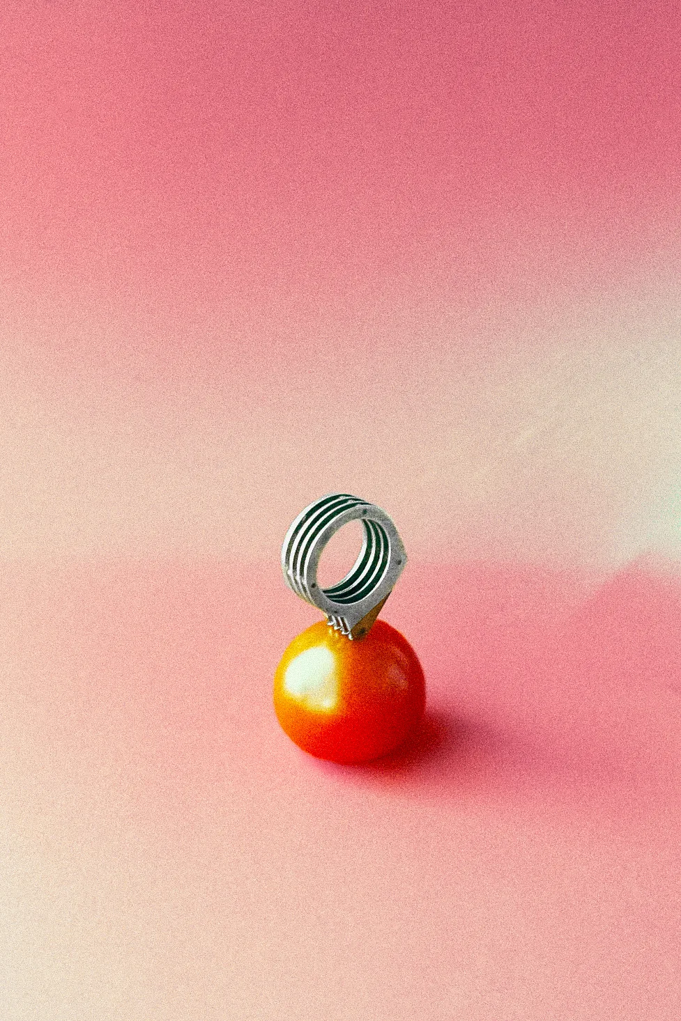 Silver and brass layered and riveted ring puncturing a cherry tomato on a pink background